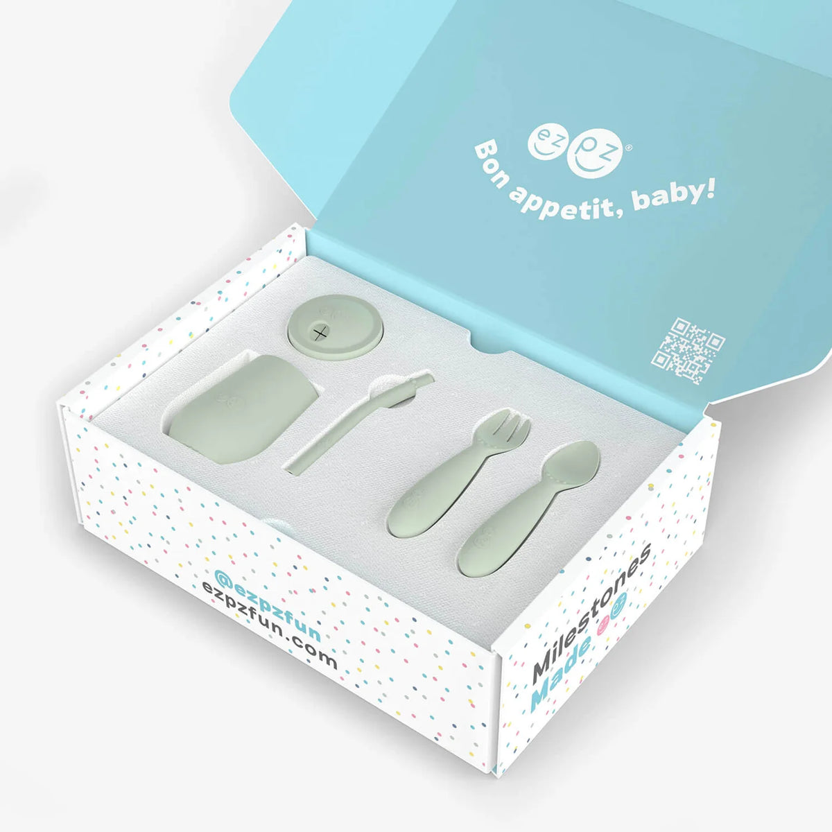 A baby feeding set in a branded gift box, including a small spoon, a fork, a round lid, and a rectangular dish, all in a pale mint green color. The open box has a light blue inner lid with the French text 'Bon appetit, baby!' and the brand name 'Milestones' visible on the side.