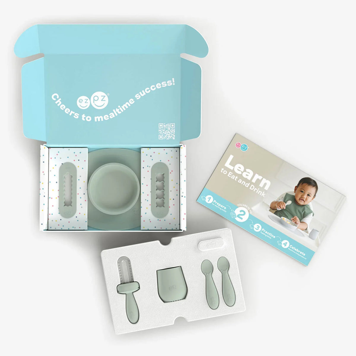 A baby feeding and mealtime success kit shown open on a white background, with a mint green bowl, silicone utensils, a branded instruction booklet, and an open light blue box labeled 'Cheers to mealtime success!'.