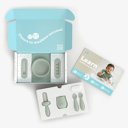 A baby feeding and mealtime success kit shown open on a white background, with a mint green bowl, silicone utensils, a branded instruction booklet, and an open light blue box labeled 'Cheers to mealtime success!'.