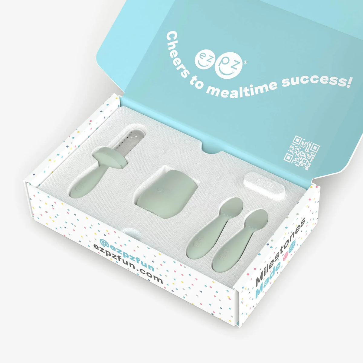 An open branded gift box containing a set of pale mint-green baby feeding tools, including a bottle brush, scoop, spoon, and a square insert. The box lid is light blue with white text reading 'Cheers to mealtime success!' and the ezpzfun logo.