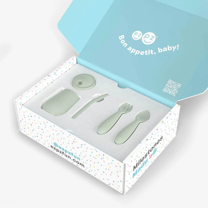A baby feeding set in a branded gift box, including a small spoon, a fork, a round lid, and a rectangular dish, all in a pale mint green color. The open box has a light blue inner lid with the French text 'Bon appetit, baby!' and the brand name 'Milestones' visible on the side.